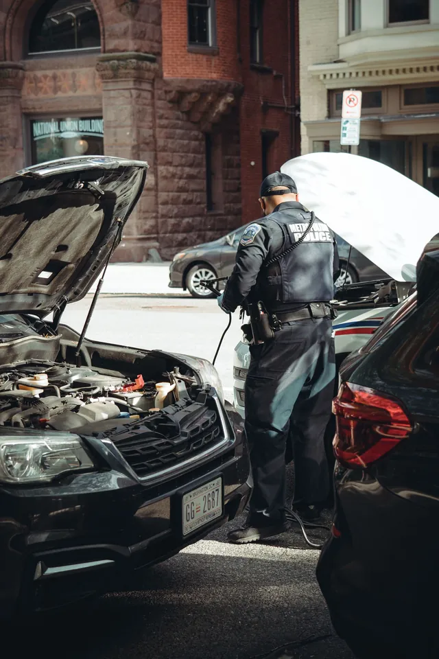 An officer connecting jumper cables between two cars with their hoods up on a city street