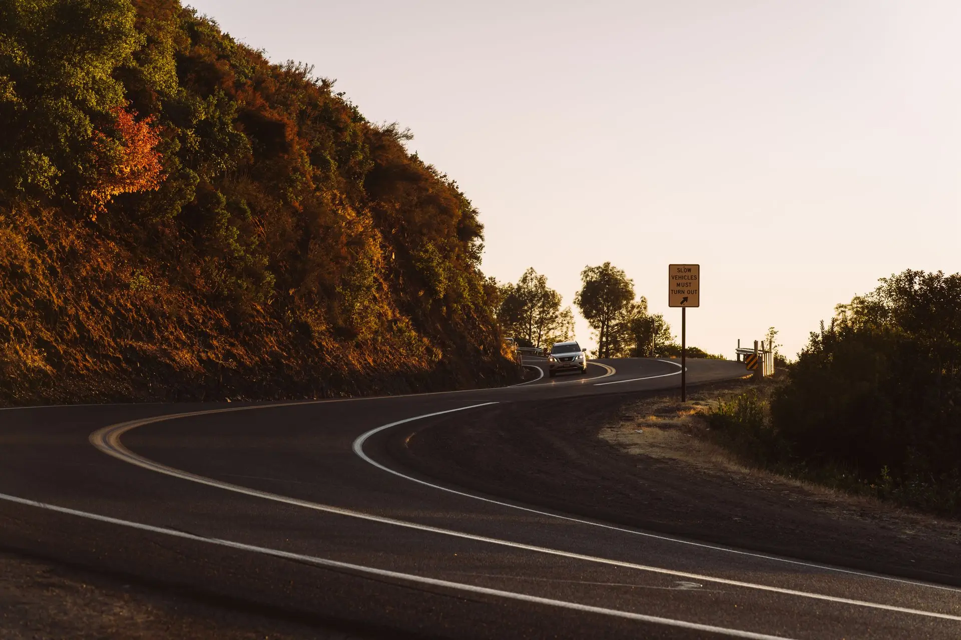 A two-lane road curving past a hillside at dusk, with a car's headlights coming around the bend